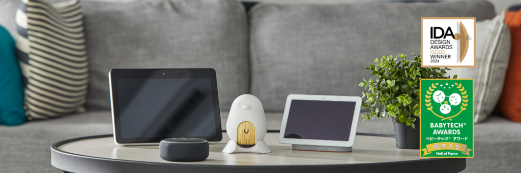 CuboAi smart baby monitor positioned on a coffee table next to smart home AI assistants including an Amazon Echo Show, Echo Dot, and Google Nest Hub. On the right, gold IDA Design Awards and green BabyTech Awards Hall of Fame badges are displayed.
